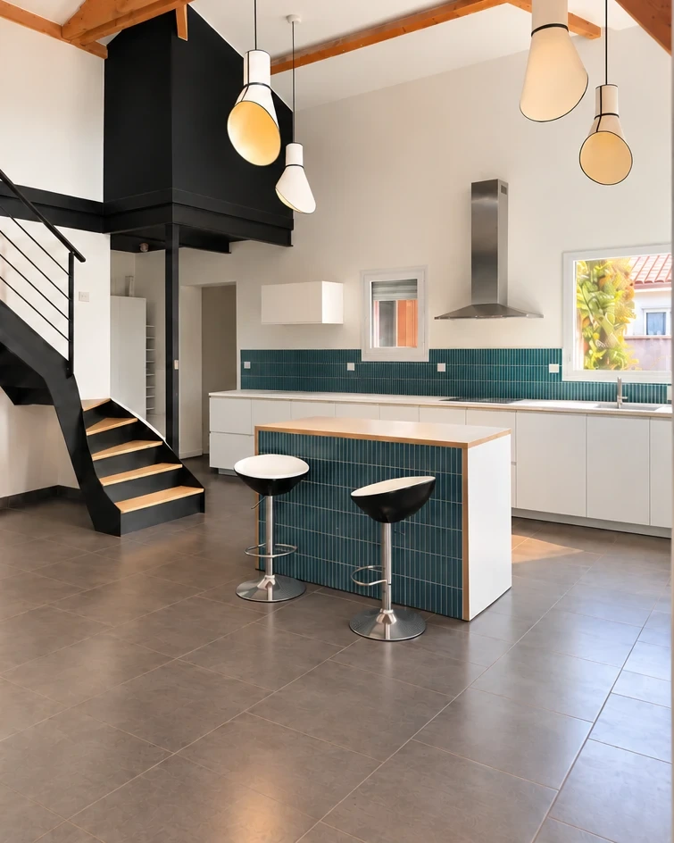 Grande pièce de vie sous charpente en bois, escalier noir, cuisine blanche et îlot carrelé de bleu, suspensions blanches.