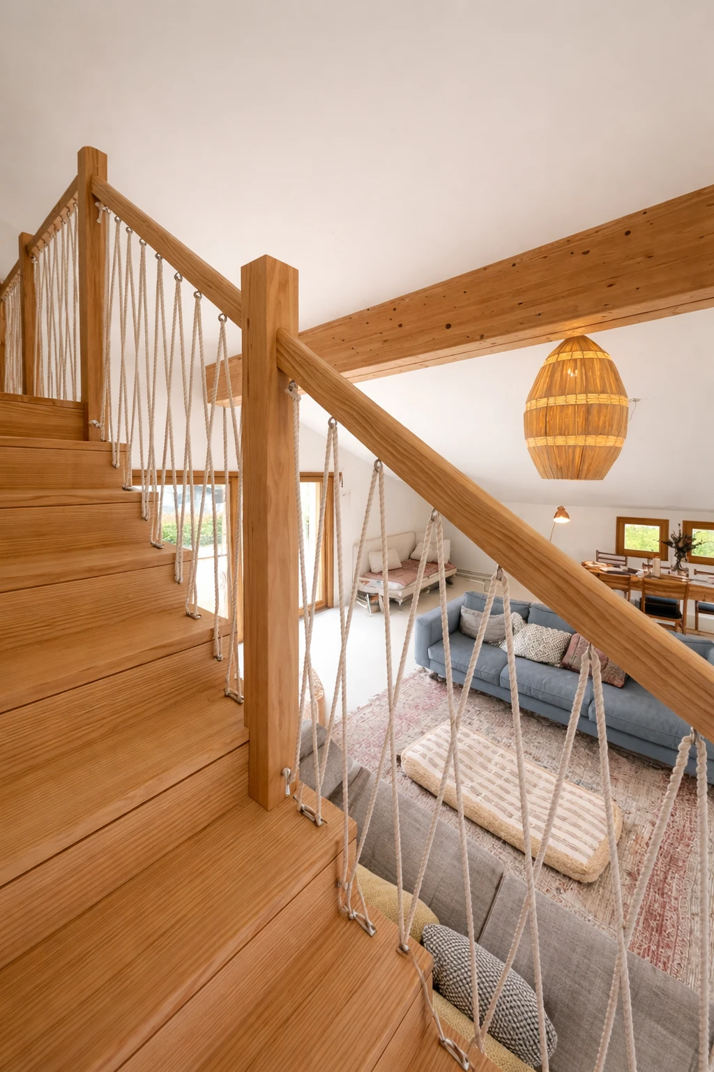Vue depuis l'escalier en bois aux garde-corps en cordage croisé, surplombant le séjour avec ses canapés bleu et gris, une suspension en rotin sous une poutre en bois et la salle à manger au fond.