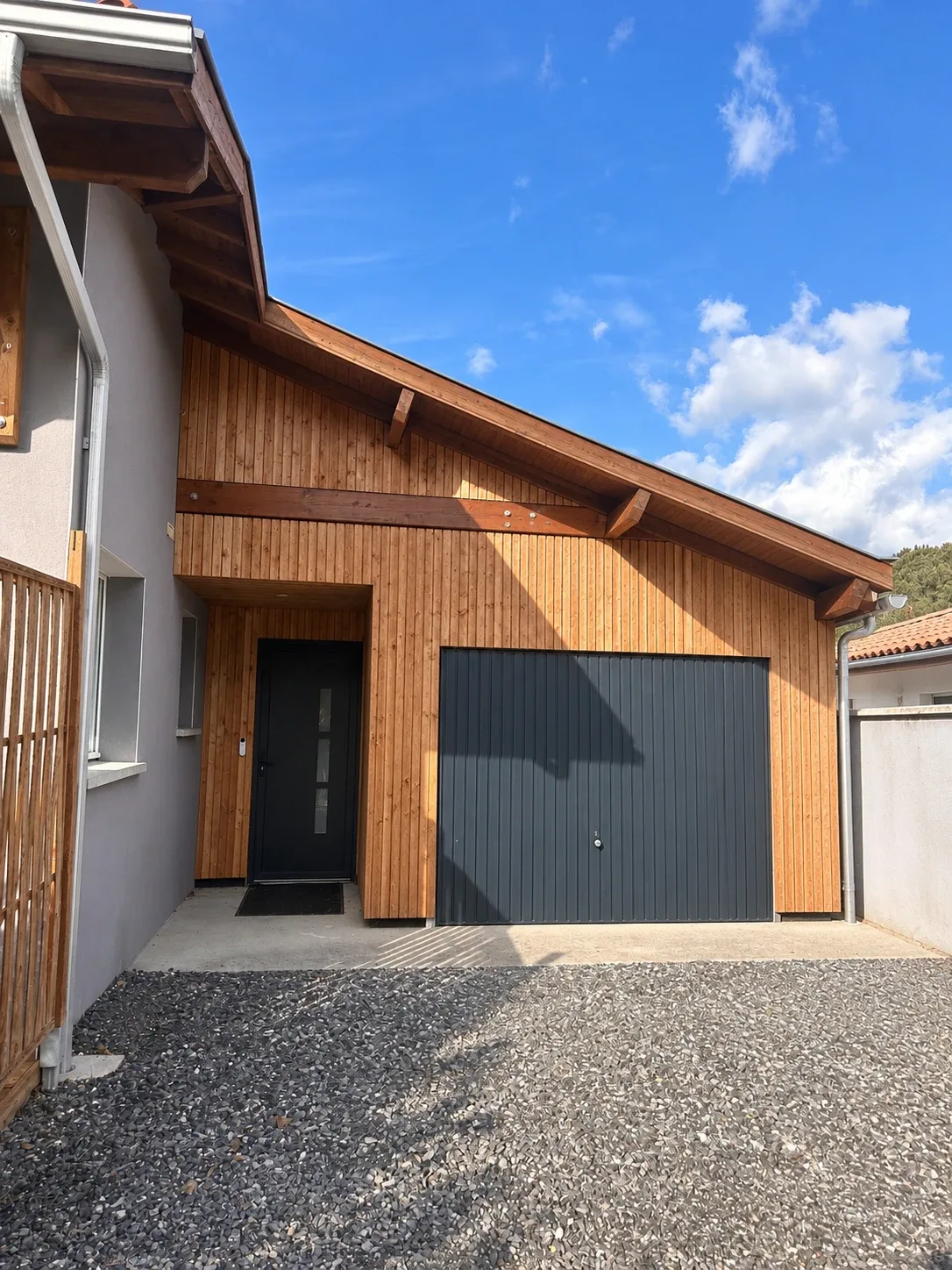 Façade de l'extension vue de face, bardage bois vertical sous la charpente apparente, porte d'entrée noire en retrait et porte de garage anthracite, entre le mur enduit gris de la maison et le mur de limite, devant une cour en gravier.