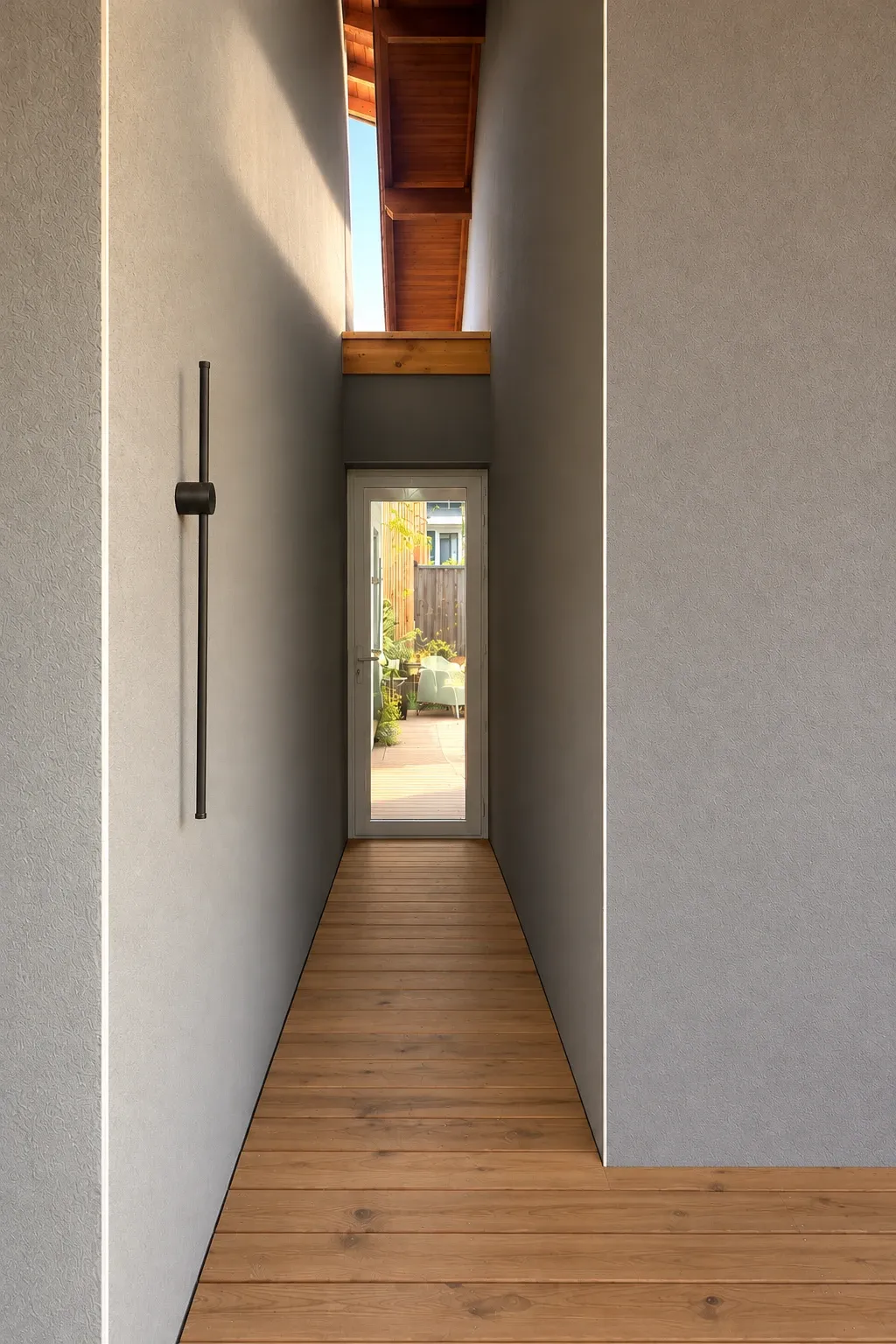 Passage étroit entre deux murs gris, plancher de bois menant à une porte vitrée ouverte sur un jardin, sous une bande de ciel